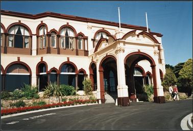 The building shows the front portico two storey Spanish influence, white stucco with red detail two figures are leaving the entrance