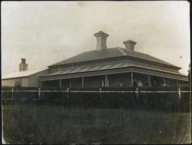 Front view of ‘Baroona’ showing family standing to the right on the verandah two chimneys, attached, weatherboard kitchen(?) to the left of the building and chimney front fence and hedge