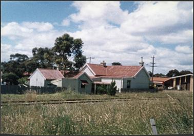 A rear view of the railway cottage, showing a white house with red iron roof, 2 chimneys, one with pot
and also outhouses the railway line visible in foreground