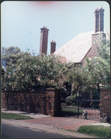 This is an Old English or neo-Tudor style clinker brick and shingle-tiled house the house has a steeply hipped main roof, showing three chimneys attached to the front and side elevations brick fence with metal spiked gate in front of the house