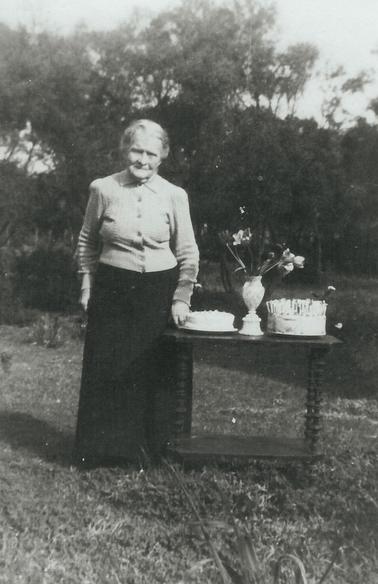 Mary Agnes Cain standing beside a table with 2 birthday cakes, one with many candles.  Vase of flowers in between the two cakes. Mrs Cain is wearing a long black skirt, light coloured blouse and long sleeved buttoned-up cardigan.