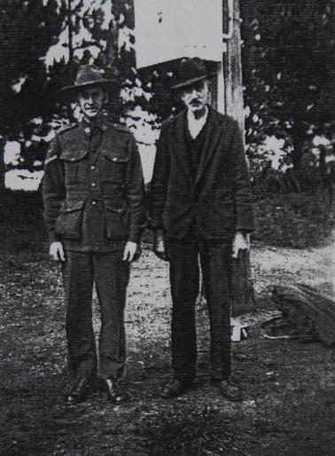 Two men - one in Army uniform, other in suit and hat, standing in front of a power pole.