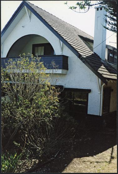 A right side view of the suburban house named ‘Rhonda’ with cypress trees partially obscuring the front and balcony of the house