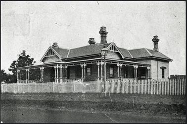 Verandah on two sides with wooden posts decorated with a fleur de lis. hipped roof, finials on peak, 4 chimneys with decoration. picket fence