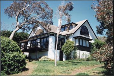 A front view of ‘Rhonda’ which is Spanish styled, white stuccoed with black trim hipped roof, two storyed with attic, balconies to front and rear, side verandah raised on left, eucalypts in garden