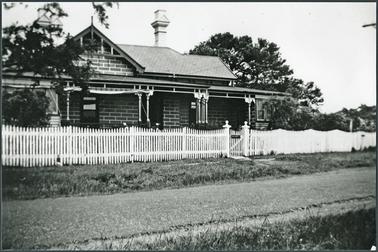 Front view, brick, verandah across front and returning wooden posts with decorative pattern, picket fence with gate posts with ornamental top, wooden filled in verandah, room on right hand side, appears to be a name plaque on gate