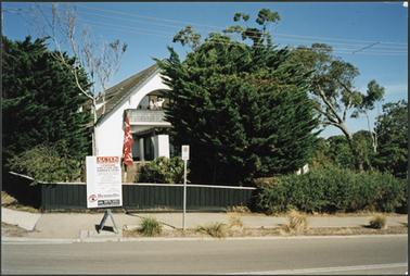 ‘Rhonda’, a Spanish styled, 2 storied home with hipped roof, white stuccoed and a balcony in front partially obscured by cypress trees in the garden a wooden fence and a ‘For Sale’ sign in front of the house