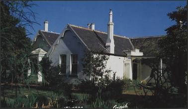 Calendar photo of Vicarage building of 1864, showing front entrance white painted stucco building, triple fronted, hipped roof with bay window on one section, patterned wooden barge boards, 4 tall chimneys, raised portico over front door, covered verandah to front door, garden view