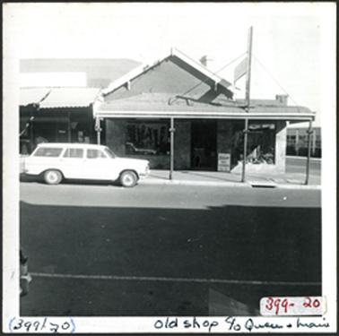 A hipped roof brick building with verandah, corrugated iron roof, on left another shop with verandah, pole with shop sign, two large windows with crazy stone surround, on right what appears to be a 1950s façade  incorporated into building Shire offices on right