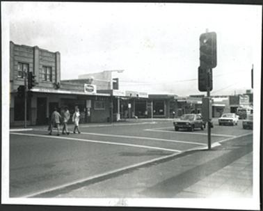 Three pedestrians on crossing two cars on road, one car parked, two story building on left one shop "Jikkys Frocks"