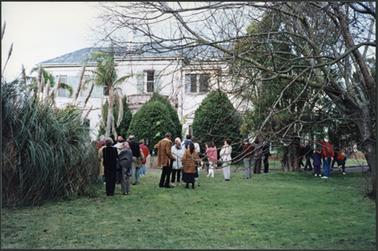 The image shows a two storied stuccoed mansion with portico, mature plantings in the front garden
