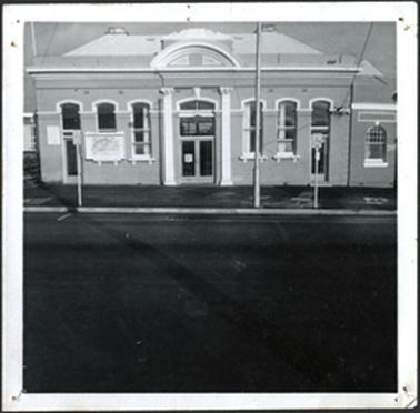 This image shows a building with arched windows on either side of glass doors which has a sign above at each end of the Mechanics building windows have been made into doorways columns on either side of centre doorway