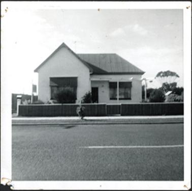 A double fronted stucco brick residence with corrugated iron roof, gabled and hipped at the front low wooden front fence