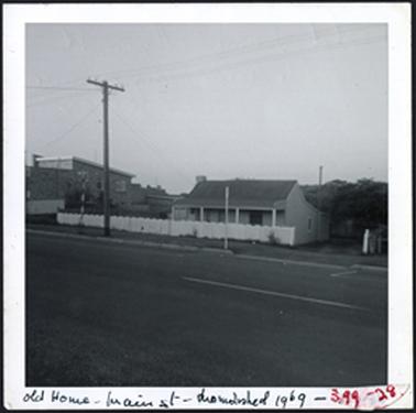 A hipped roof residence with verandah which has four posts, corrugated iron roof, rendered brick white picket fence weather board addition on rear to storey 1950s building on left