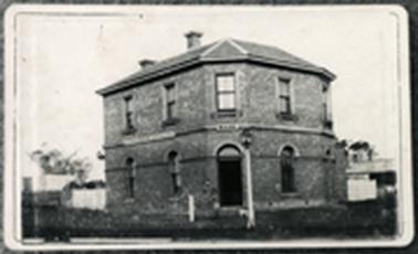 A two storey corner brick building five upper windows four lower windows and doorway on corner with arches  two chimneys fence and street lamp in front smaller buildings to left and right altered over time