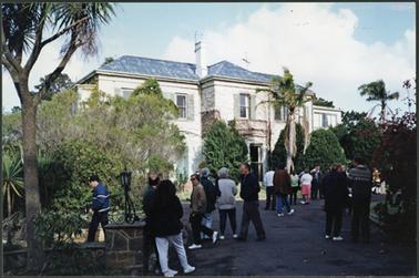 ‘Craigie Lea’  the image shows a two storey brick stucco Italianate mansion, one chimney and shutters on the windows mature trees, photo taken auction day and shows people gathering