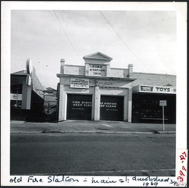 A brick stuccoed building with two entrances, parapet above with central detail and two end details, Fire Station 1919 on parapet, ‘For Sale’ notice, alleyway used for fire brigade practice, with a bell tower, on left, two storey Mornington Car Sales Yard, on right Toy and Hobby Shop