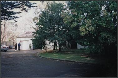 A view through the garden with mature trees to the garage which is single storey, one section with door and centre open, winding driveway