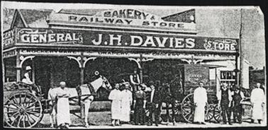 The Railway Store and Bakery advertising on the building states that it is a General Store owned by J.H. Davies. Other advertising is for the sale of drapery, crockey ware, grocery, boots and shoes. A building with a hipped roof, posts with plain ornamentation. In the front of the building are two horses and carts and another which is a bakers delivery cart. There are also ten adults and one child in the picture
