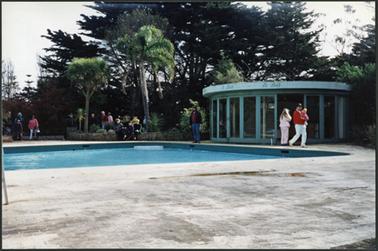 The image shows the rectangular pool with circular bar structure and full length doors, all opening, mature cypress trees in background 