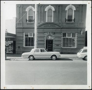 A two storey stone building, with three windows in top storey with double arched lintels and floral design central stone arch over false 'porch' entrance, a arched doorway with stone work surround picked out large square windows Coles variety store on left opposite Albert Street