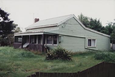 Depicts an old weather board house painted light green with white trim around the windows. The photo has been taken side on. There is a verandah along the front of the house with a brown lattice railing. The grass in the front and side garden is overgrown with a wooden pailing fence in the foreground.