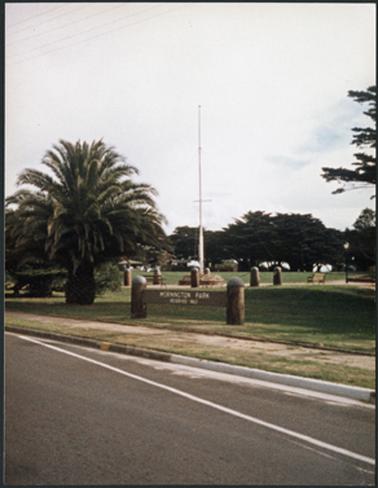 The flagpole erected in the Mornington Park surrounded by palm and other mature trees. In the foreground 
is a sign “Mornington Park”