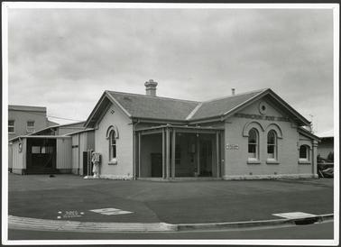 A front view of the painted brick Post Office building with a slate roof and corner wooden verandah or portico, the old styled weighing machine is situated out the front of the building on the Main Street side, the corrugated tin sheds used by the Post Office can be seen at the rear of the building 
