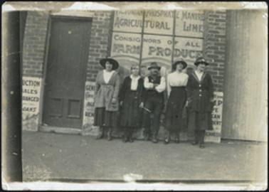 Building showing brick uprights, wooden front door four women and one man outside building building advertising ‘Auctions, Agricultural Lines, Farm Products and AMP Insurance’