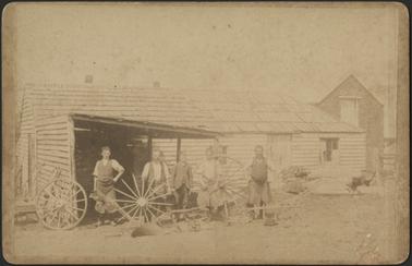 Image of five men standing outside a wooden building a wooden slatted building with slanted shingled roof
and loft to the right 