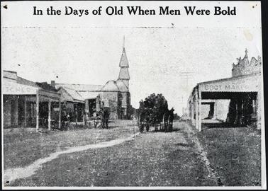 Black and white image of the Main Street of Mornington, with two drays on the road, one containing the local football team to the left of the image are shops, one a Butchers and The Grand Hotel on the right is Bartletts 
the Bootmaker, a quote on the photo which reads “In the Days of Old When Men Were Bold”