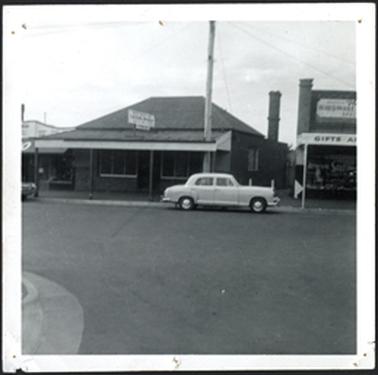 The image is of three shops, Peninsula Radio on left, hipped roof with verandah centre and the shop on the
right has a parapet and verandah walkway between shops a car in front of the buildings