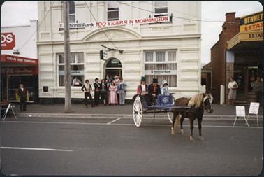 View in Main Street, Mornington. The image is of a horse and carriage with one woman and two men dressed in Victorian costumes in front of the National Bank. A group of people in Victorian costumes are standing in the doorway of the National Bank. A poster advertising ‘National Bank 100 Years in Mornington’ is on the National Bank building. This image was taken in 1988 on Australia Day