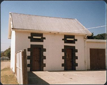 A stucco building with a hip roof and bluestone surrounds to the doors and vents above, garage? at right side
of the building