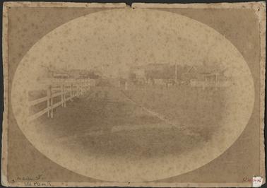 A very faded image of the Main Street looking south a wooden fence shown on the left and the Post Office building on the right a horse and buggy outside the Post Office the image is oval shaped and backed onto cardboard