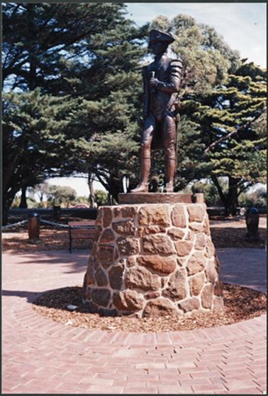 A bronze statue of Matthew Flinders which is on top of brick work surrounded by a paved area with mature trees in the background and is located in the Mornington Park, the Esplanade, Mornington