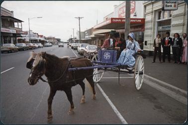 View down Main Street, Mornington the image is of a horse and carriage with one woman and two men dressed in Victorian costumes a group of people in period costume are standing outside the National Bank this image was taken in 1988 on Australia Day