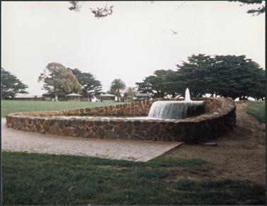 A stone water feature (fountain) surrounded by a paved area there are trees and pavilions in the background