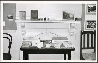 A view of the historical society museum display in 1970 showing the fireplace with a wooden table placed in front displaying minerals or fossils