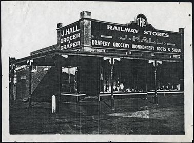 View on the corner of Blake and Main Streets Mornington the image is of a shop front with verandah advertising on front of the building - ‘The Railway Stores - J. Hall, Drapery, Grocery, Ironmongery, Boots and Shoes’