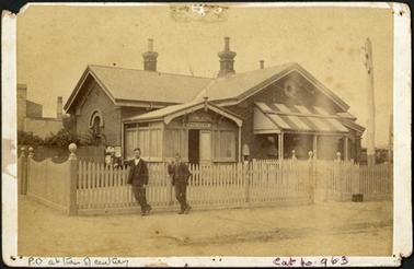 A front view of the Post Office building, with an enclosed entrance a picket fence is built around the Post Office two men in period clothing are leaning against the fence the photograph is mounted onto card