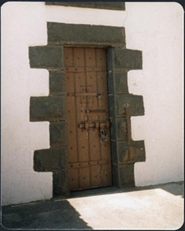 The image shows a heavy wooden door with bolts and locks stucco building with blue stone door surrounds