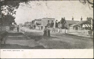 A view looking up the Main Street, Mornington, from the Esplanade, the Old Post Office Building is shown on the right two women dressed in Edwardian clothes with hats are featured standing on the road near the Post Office