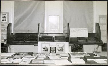 A view of the historical society museum display in 1970 showing a range of photographs, books and other literature which have been set out on the Post Office counter and writing desks