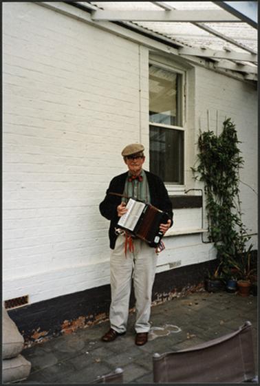 Photo of Mac David son Vice President, at the Garden Party standing in front of  window at The Vicarage Ross St. November 1994, holding an accordion

Query on date noted 1992 and on photo 1994