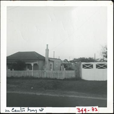 A side view of house with verandah, hipped roof, chimney and picket fence