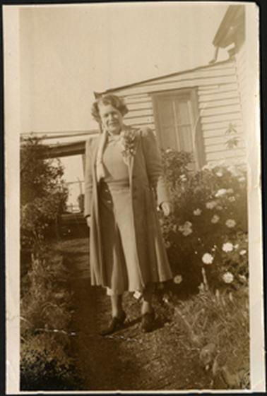 The image shows Wyn Bradford standing on a garden pathway with part of a wooden building behind her