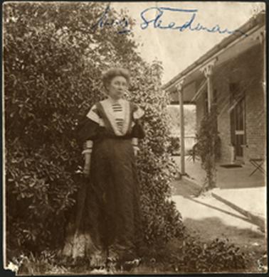 The image is of Mrs Steedman photographed standing in front of shrubbery, part of a house can be seen at the right of the photo