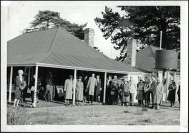 The image is of a hipped roof building, with verandah and two chimneys, it appears an extended building to right of photo, a water tank in front. Several people standing under verandah and in front of the building