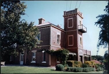 A side view of Sutton Grange, the house is two storey with a one level concave roof, verandah abutting the square tower the main roof is hipped, established lawns and gardens